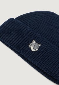 Zīda adīta cepure tumši zilā krāsā ar ribotu tekstūru; priekšpusē ir izšūta lapsas galvas emblēma baltā un pelēkā krāsā.
