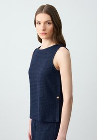 Débardeur sans manches bleu marine en tissu texturé, avec encolure arrondie et logo doré sur le côté. Design à coupe droite.