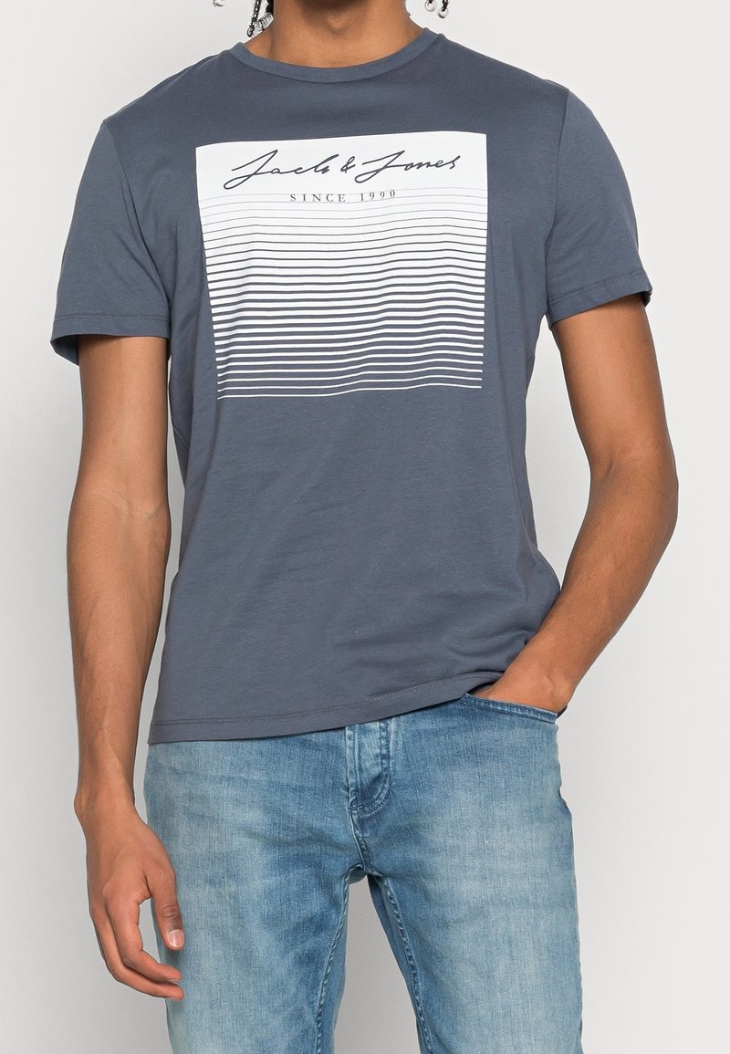 T-shirt en coton gris à manches courtes, avec un graphique rectangulaire blanc comportant des rayures bleues et blanches alternées ainsi que du texte.