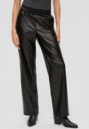 Vrouw die zwarte brede nepleren broek draagt, zwarte laarzen met hoge hak en spits toelopende neus, en een donker metallic mouwloze top.