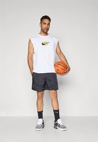 Valge varrukateta sportlik särk kollase ja musta logoga, koos mustade lühikestega. Hoides oranži korvpalli, kannab musti sokke ja tossusid.