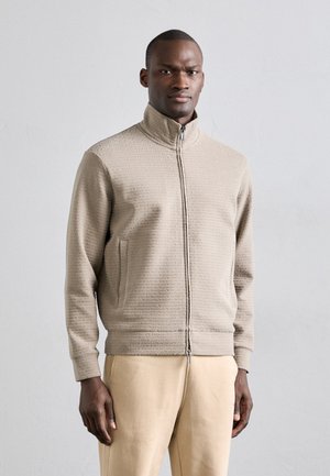 Veste zippée beige clair en matériau texturé, avec un col montant, des poches latérales et des poignets et un ourlet côtelés.
