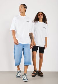 T-shirts blancs surdimensionnés avec le logo "LEGENDS", shorts en denim bleu, shorts de vélo noirs, baskets vertes et sandales noires, le tout sur un fond neutre.