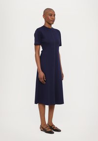 Navyblaues Kleid mit kurzen Ärmeln und einem runden Ausschnitt, ausgestattet mit einer zentralen Naht und glattem Stoff. Schwarze Riemchenschuhe mit Details.
