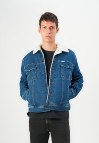 Denim jack met een lichte nepbont kraag, blauwe kleur, knoopsluiting, twee borstzakken en contrasterende stikdetails. Gedragen over een zwart shirt.