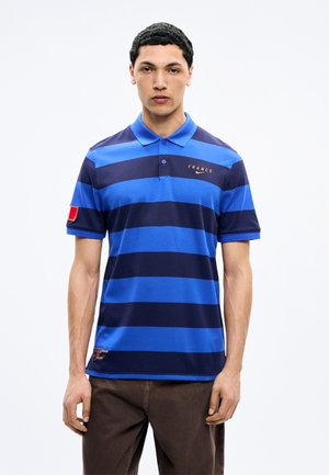 Homme portant un polo à rayures horizontales bleues et bleu marine avec l'inscription "France" et le logo Nike, debout devant un fond blanc uni.