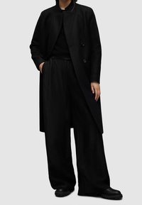 Czarny płaszcz z mieszanki wełny z wysokim kołnierzem i podwójnymi guzikami, zestawiony z luźnymi czarnymi spodniami i masywnymi czarnymi butami.