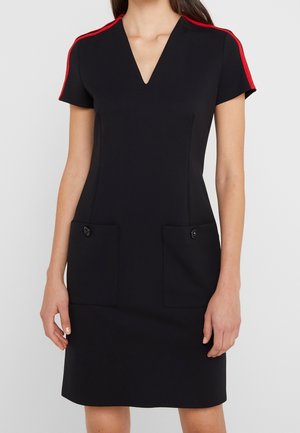 Femme portant une robe noire ajustée à manches courtes, des rayures rouges aux épaules, un décolleté en V et deux poches avant boutonnées.
