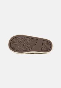 Suela de zapatilla con una base de goma marrón, que presenta un patrón texturizado para tracción y un borde circundante en blanco roto.