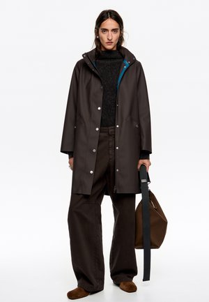 Brązowy wodoodporny płaszcz przeciwdeszczowy z zatrzaskami i kapturem, nałożony na ciemny sweter i brązowe szerokie spodnie, trzymany w torbie na ramię.