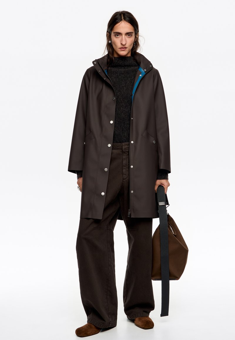 Abrigo de lluvia marrón impermeable con botones a presión y capucha, superpuesto a un suéter oscuro y pantalones anchos marrones, sostenido con un bolso de hombro.