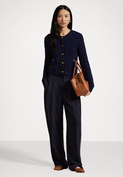 Tummansininen cable-knit neuletakki kullanapainikkeilla ja etutaskuilla, yhdistettynä tummiin leveälahkeisiin housuihin ja ruskeaan nahkatelttakassiin.