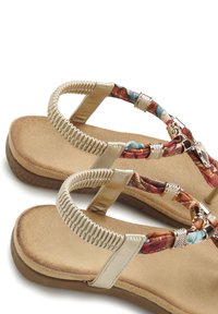 Des sandales plates beiges avec une semelle intérieure texturée, des brides à motifs floraux et des accents métalliques. Elles présentent une sangle de cheville flexible pour le confort.