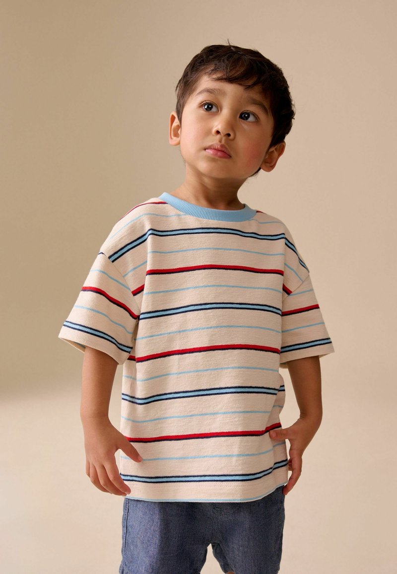 Ragazzo giovane in piedi con le mani lungo i fianchi, indossa una camicia beige a righe con linee rosse, blu e nere e pantaloncini blu su uno sfondo neutro.