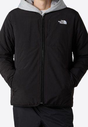 Osoba ubrana w czarną kurtkę na zamek błyskawiczny z logo The North Face i szarą bluzę z kapturem pod spodem, ręce w kieszeniach kurtki.