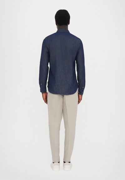 Dark blue long-sleeve shirt with a collar, back yoke, curved hem, paired with light beige trousers and white sneakers.
