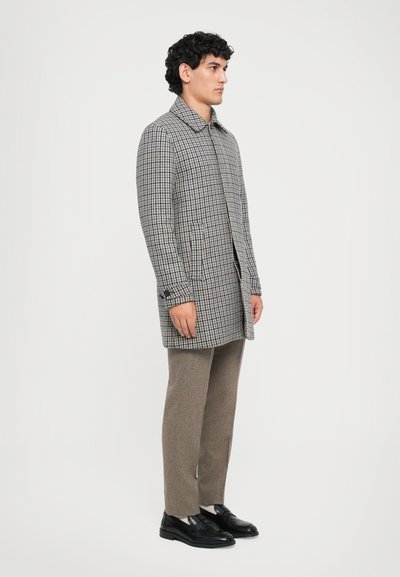 Tan- och brunt rutigt trenchcoat med krage, förlängd form, framfickor och svarta skor, ihop med ljusa byxor.