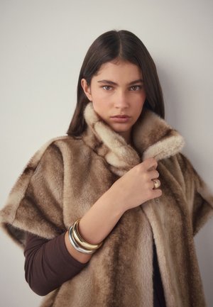 Frau mit langen dunklen Haaren trägt einen braunen Pelzmantel, hält den Kragen und ist mit Gold- und Silberarmbändern sowie einem goldenen Ring geschmückt.