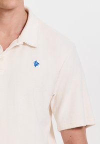 Terepmintás off-white színű, puha textúrájú pólóing, kis kék korall hímzéssel a mellrészén. Rövid ujjakkal.