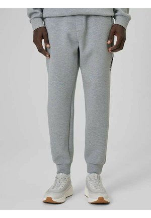 Osoba ubrana w jasnoszare spodnie dresowe i białe tenisówki, stojąca na jednolitym jasnoszarym tle.