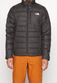 Doudoune noire avec col haut, texture matelassée, fermeture éclair devant et détail de logo sur la poitrine gauche. Portée sur un pantalon orange.