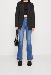 Svart dubbelknäppt blazer över en kort topp, kombinerad med ljusblå flare jeans och kraftiga svarta plattformsskor. En liten svart handväska ingår.