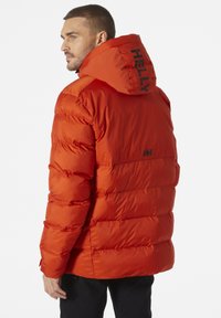 Červená péřová bunda s kapucí, bočními kapsami a černými akcenty značky. Materiál vypadá hladce a je izolovaný s prošívaným designem.