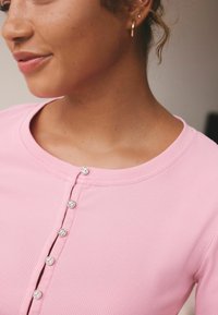 Top corto a maniche corte ribbed rosa chiaro con scollatura arrotondata e cinque bottoni a pressione metallici lungo la parte anteriore. Texture liscia, design aderente.