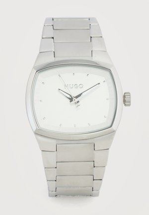 Ceas de mână din metal argintiu cu cadran pătrat, marcaje orare minimaliste și logo-ul "HUGO" pe cadranul alb, curea din zale metalice și coroană pe partea dreaptă.