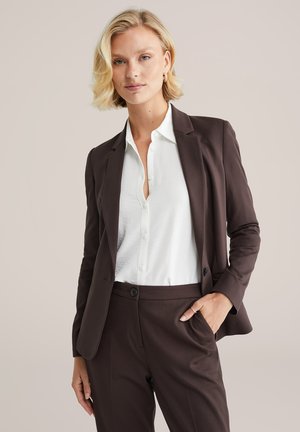 Blazer marrone su misura sopra una camicia bianca con bottoni, abbinato a pantaloni coordinati. Tessuto morbido, design aderente e dettagli discreti dei bottoni.