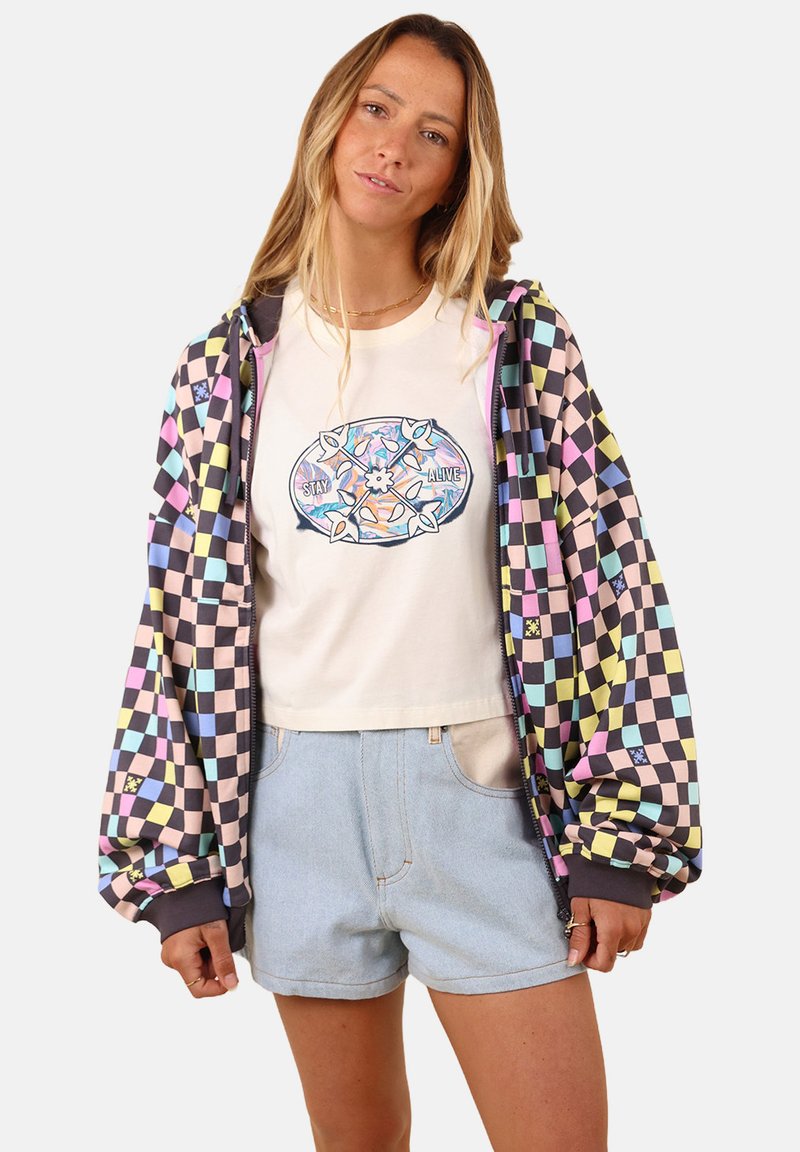 Hoodie à carreaux multicolores avec des carrés noirs, roses et bleus ; tee-shirt graphique blanc avec "STAY ALIVE" ; shorts en denim clair.