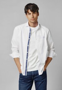 Witte button-up shirt met een blauwe accent langs de knoopsclosure, gecombineerd met een witte crew neck t-shirt en donkere spijkerbroek. Soepele textuur.