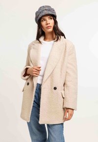 Žena v textúrovanom krémovom kabáte cez bielu košeľu a modré džínsy, doplnená o farebnú textúrovanú čiapku.