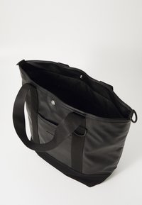 Sac fourre-tout noir en cuir lisse avec deux poignées extérieures, fermeture à pression en haut, poche intérieure et base en tissu.