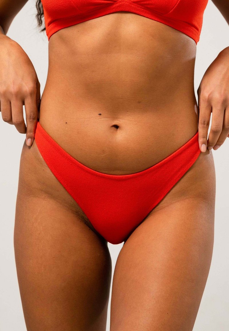 Röd bikiniunderdel med högskuren design, slät textur och minimal täckning, med kontrasterande sömmar längs kanterna.