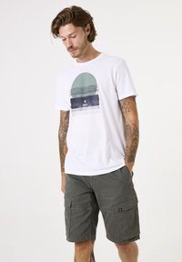 Camiseta blanca de algodón con un gráfico circular en verde y azul, combinada con pantalones cortos de cargo en verde oliva con bolsillos y un parche con logo.