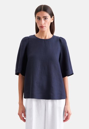 Kobieta o prostych ciemnych włosach, ubrana w luźną granatową bluzkę z krótkim rękawem i białą spódnicę, stojąca na jednolitym tle.