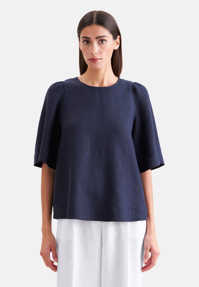 Femme aux cheveux raides et foncés portant un chemisier ample bleu marine à manches courtes et une jupe blanche, debout devant un fond uni.