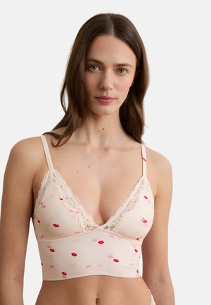 Frau mit langen braunen Haaren, die ein hellrosa Spitzen-Bralette mit roten und pinken Lippenabdrücken auf weißem Hintergrund trägt.