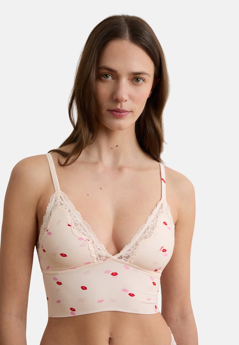 Femme aux longs cheveux bruns portant un bralette rose clair bordé de dentelle avec un motif de lèvres rouges et roses sur fond blanc.