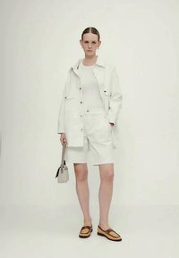 Balta denima jaka un šorti kopā ar baltu topu, brūniem loafers un mazu beigu krāsas somiņu. Gludas tekstūras un minimālistisks dizains.