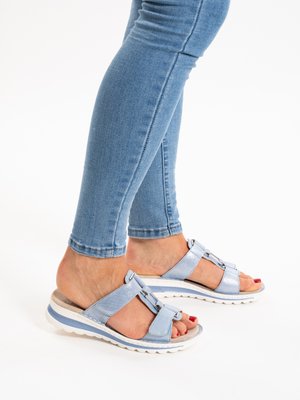 Persoon draagt lichtblauwe metalen instapsandalen met dikke witte zolen en een blauwe spijkerbroek, staand tegen een witte achtergrond.