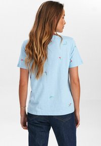 Camiseta de algodón azul claro con mangas cortas, decorada con pequeñas flores bordadas en rosa y verde. Corte relajado con cuello redondo.