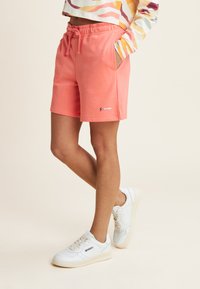Pantalones cortos coral con cintura elástica y bolsillos laterales, combinados con zapatillas blancas que tienen un logo. Textura suave y ajuste casual.