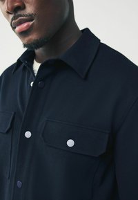Primer plano de un hombre que lleva una camisa de botones azul marino oscuro con bolsillos con solapa y botones a presión blancos.