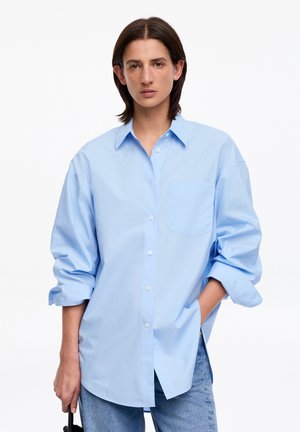 Person trägt ein übergroßes hellblaues Hemd mit aufgekrempelten Ärmeln und blaue Jeans, steht mit einer Hand in der Tasche und hält eine schwarze Tasche.