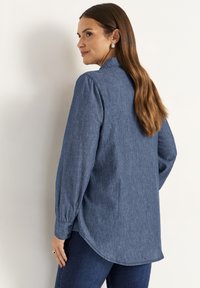 Denim blue, long-sleeved shirt with a rounded hem, buttoned cuffs, and a subtle texture, worn with dark jeans. Back view.