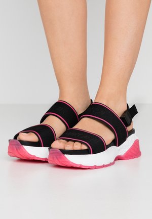 Fødder iført sorte sporty sandaler med pink kant og tyk hvid sål med pink mønster, vist mod en ensfarvet grå baggrund.