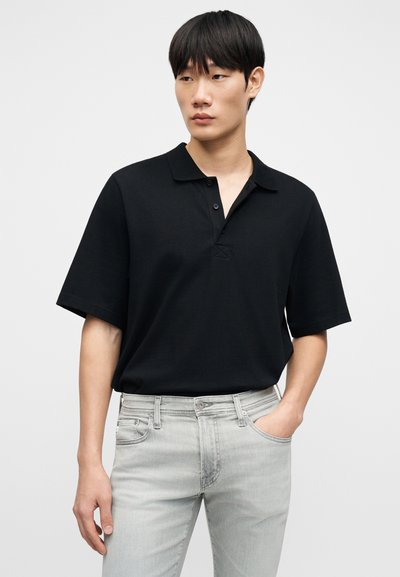 Μαύρο πόλο μπλουζάκι με γιακά και τρία κουμπιά, κατασκευασμένο από μαλακό ύφασμα, συνδυασμένο με ανοιχτό γκρι τζιν. Διαθέτει άνετη εφαρμογή και κοντά μανίκια.