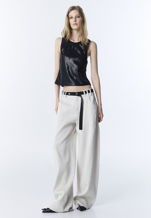 Große Frau trägt ein ärmelloses schwarzes Paillettenoberteil, weite weiße Hosen mit einem schwarzen Gürtel und schwarze spitze Schuhe, steht vor einem weißen Hintergrund.